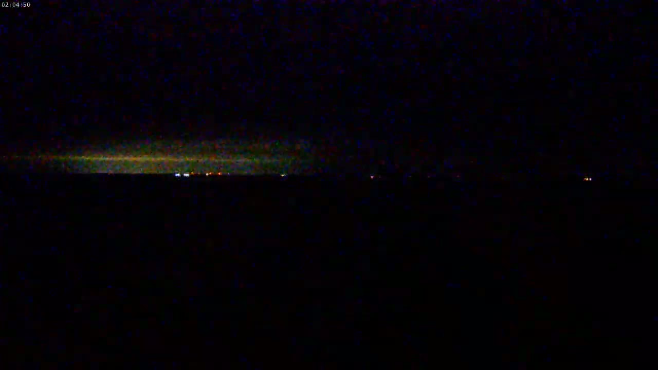 A dark, nighttime landscape shows a faint, illuminated horizon line with a few distant lights under a mostly clear sky.