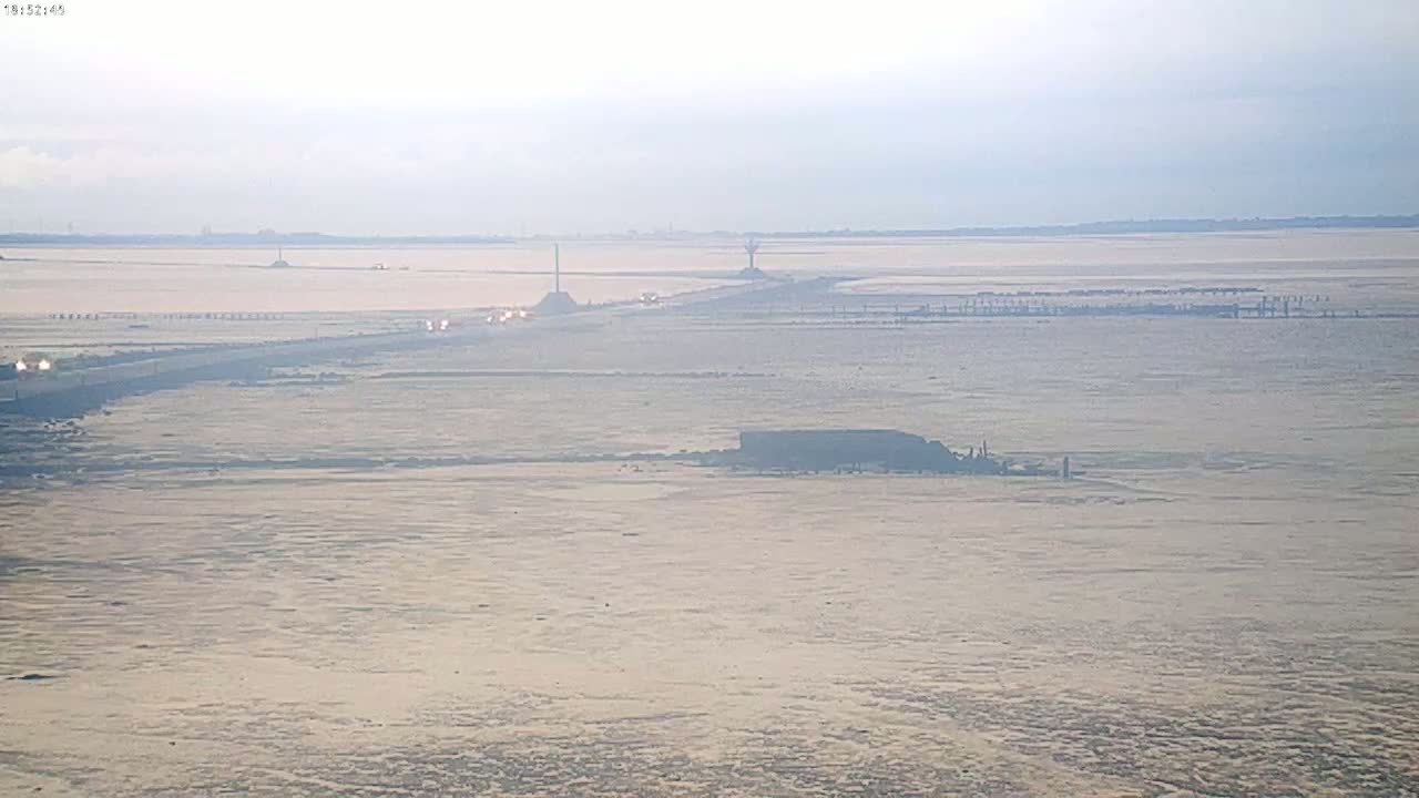 A long, flat expanse of light-brown land stretches to the horizon under a hazy sky, with a road and sparse vehicles visible in the midground.