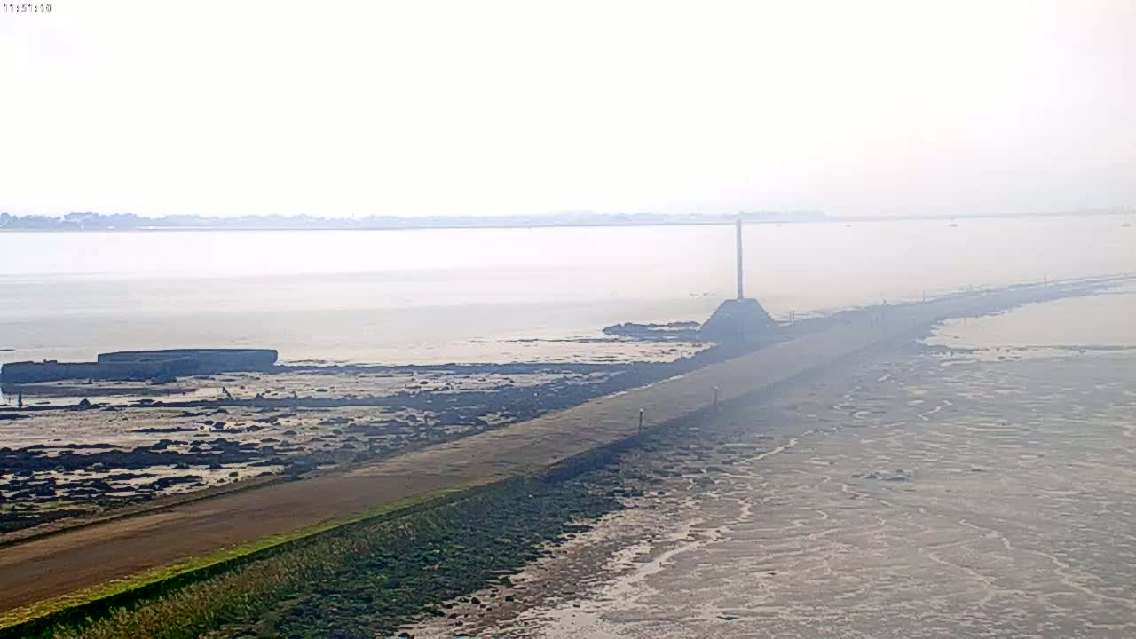 A long, paved causeway extends into a hazy, calm body of water, with low-lying land and a tall pole visible in the distance.