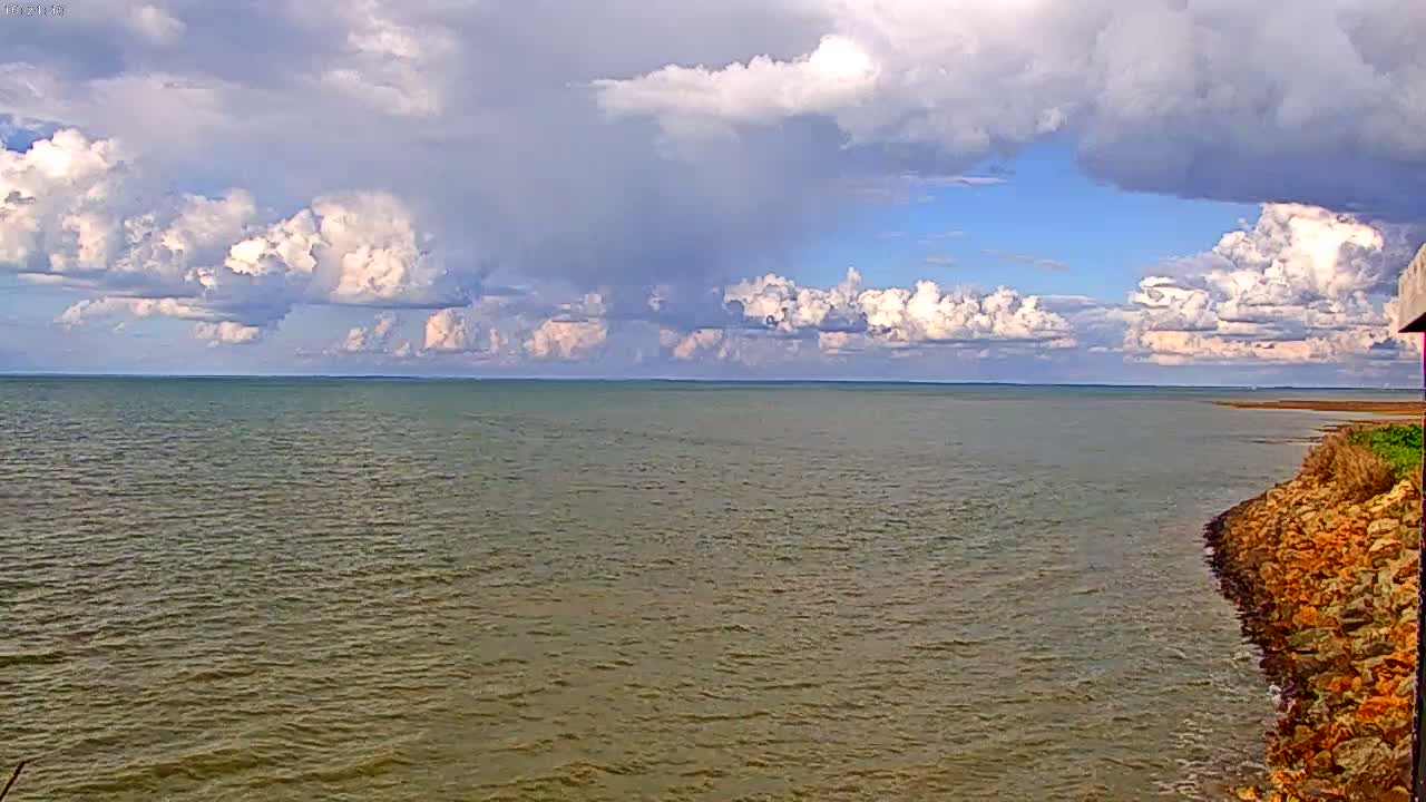 A partly cloudy sky overlooks a calm body of water with a rocky shoreline to the right.