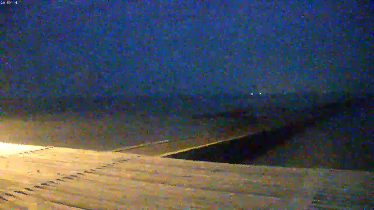 A dark, nighttime scene shows a snow-covered, sloped surface leading down to a dark body of water under a dark blue sky.