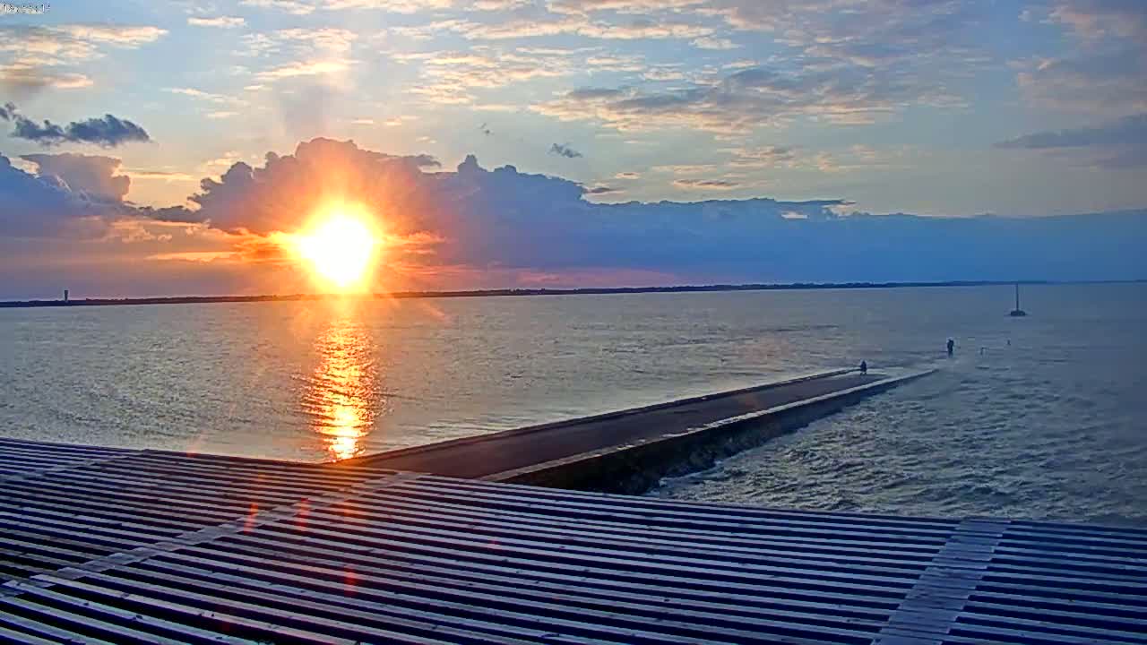 A sunset over a calm body of water, viewed from a slatted surface, shows a long, thin road extending into the water.