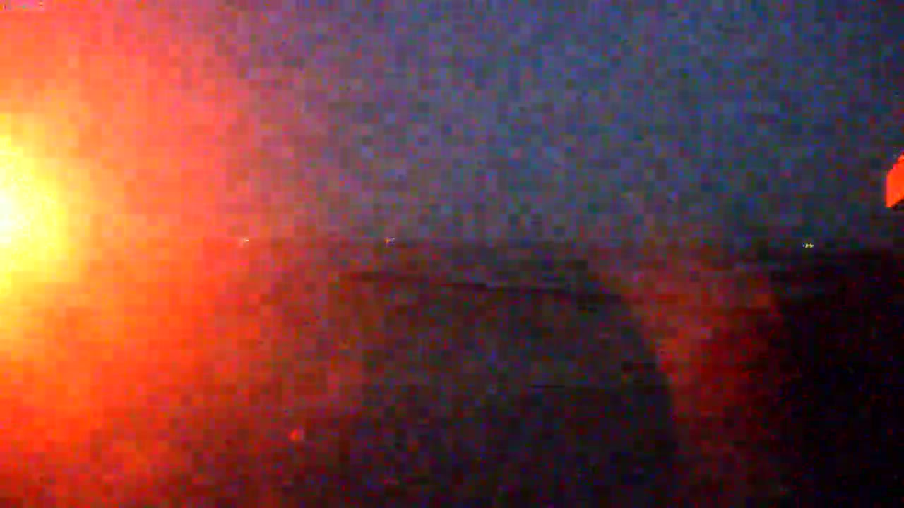 A large, bright orange light, possibly a fire or explosion, dominates the left side of a dark, nighttime scene.