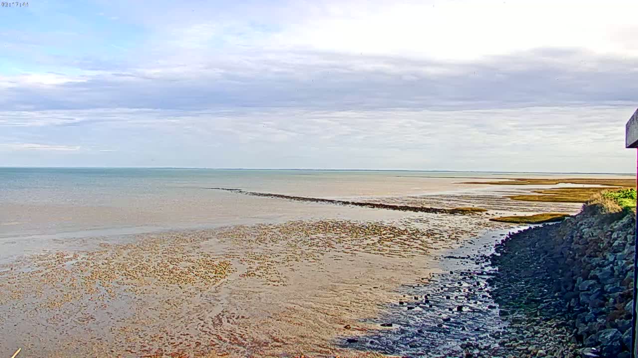 A mostly cloudy sky overlooks a wide expanse of shallow, muddy water receding from a rocky shoreline.