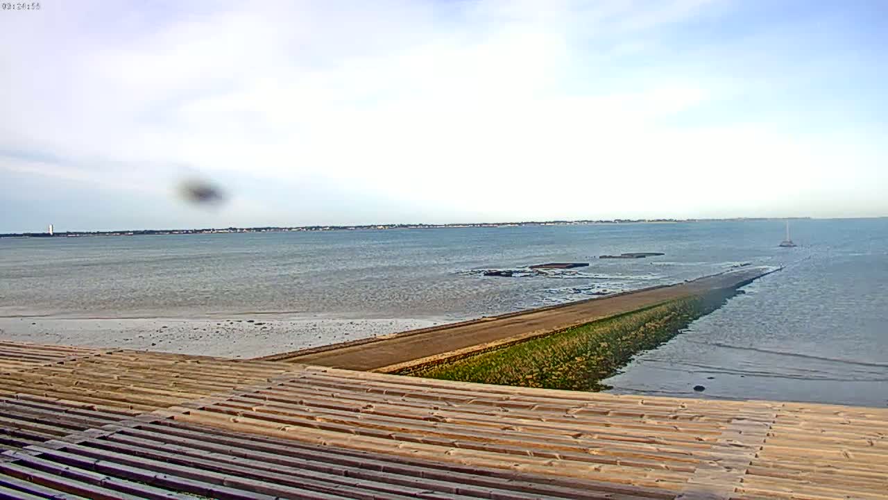 A long, narrow causeway extends from a wooden structure into a calm, light-blue sea under a mostly cloudy sky.