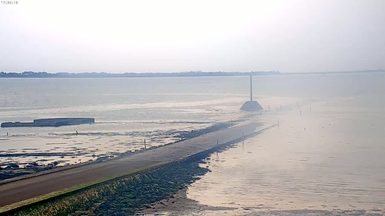 A hazy view of a long, narrow road extending into a calm body of water, with a distant shoreline visible in the background.