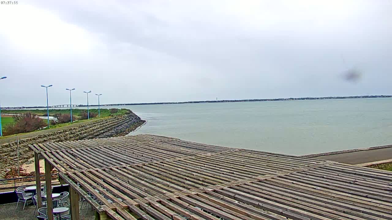 A wooden pergola overlooks a calm body of water under an overcast sky, with a distant bridge visible on the horizon.