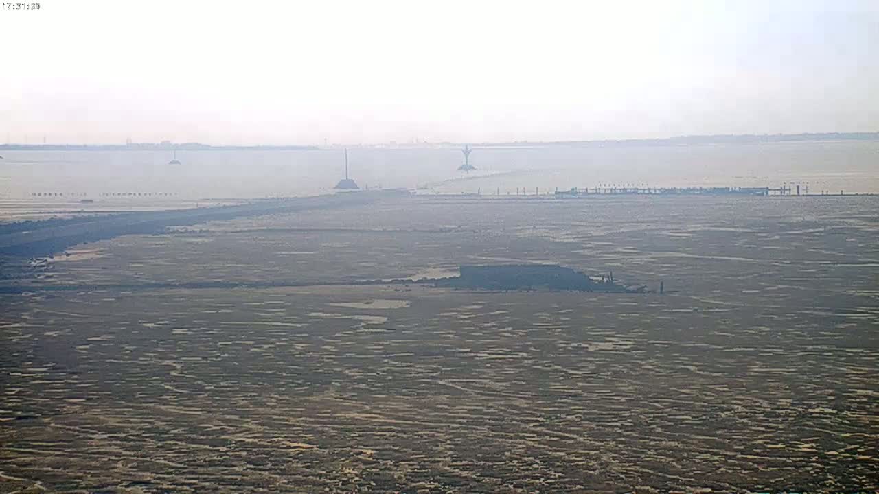 A hazy, flat expanse of land and water stretches to the horizon, featuring several tall structures in the distance.