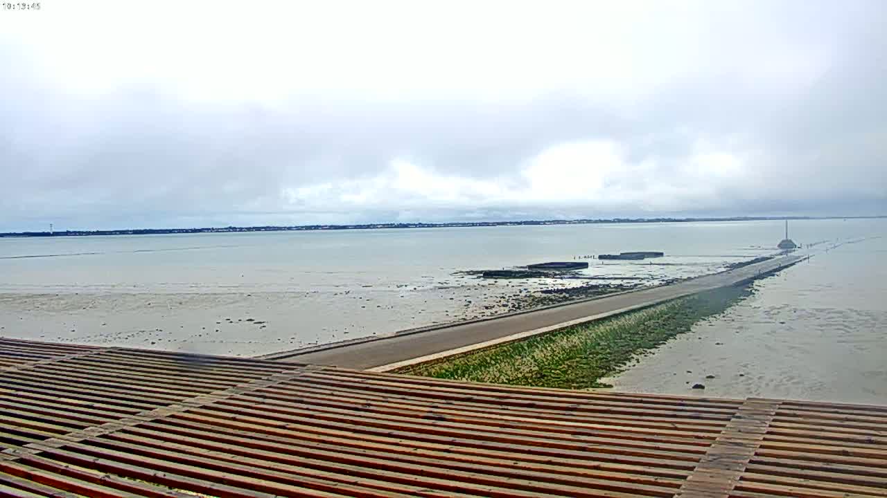 A long causeway extends into a calm, overcast body of water, receding to reveal mudflats, with a wooden slatted structure in the foreground.