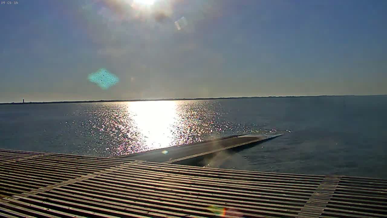 A wooden slatted platform extends over a calm body of water reflecting the bright sun on a clear day.