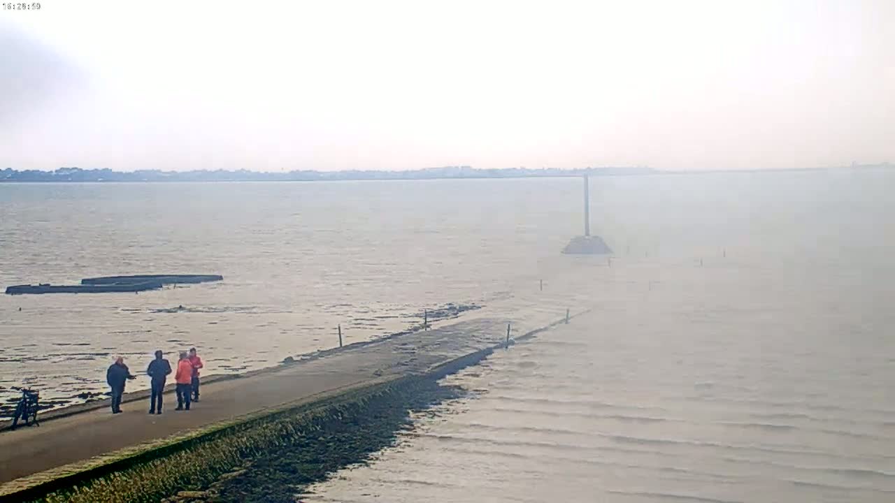 A hazy, overcast day shows several people walking on a long, narrow road that extends into a calm body of water, with submerged structures visible in the distance.