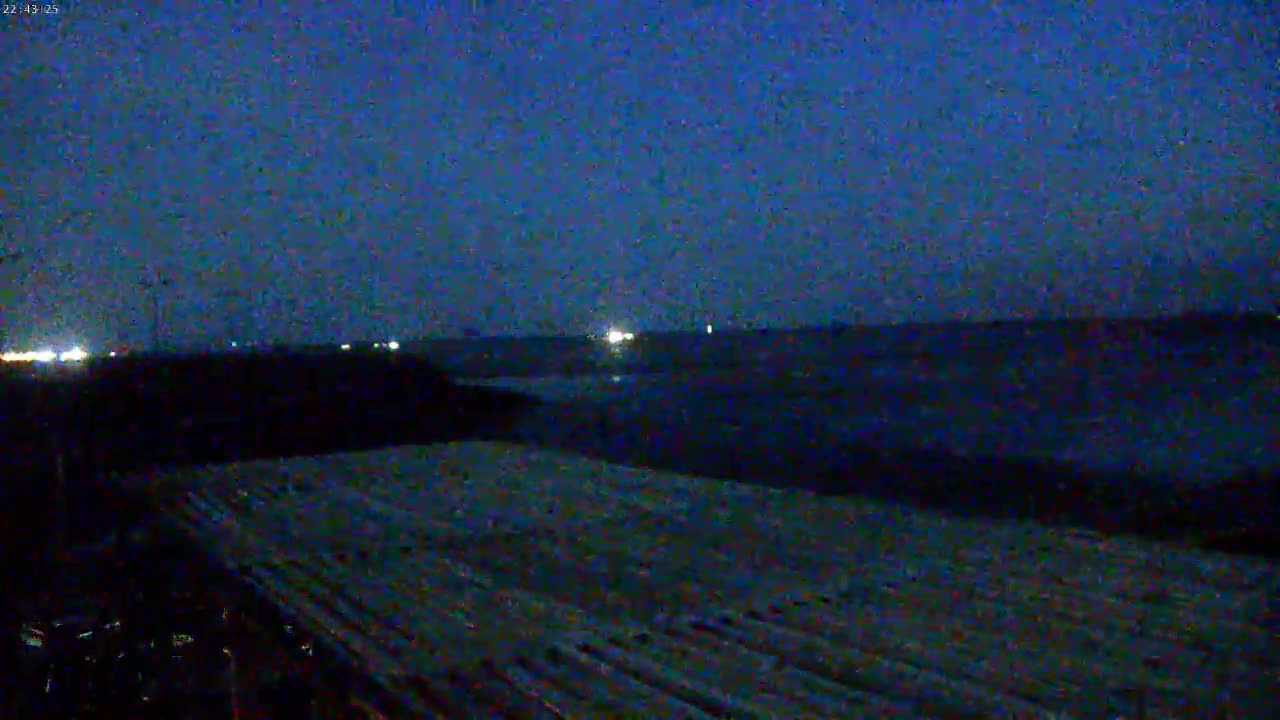 A dark, nighttime scene shows a flat, textured surface in the foreground, with a dark body of water and distant lights under a twilight sky.
