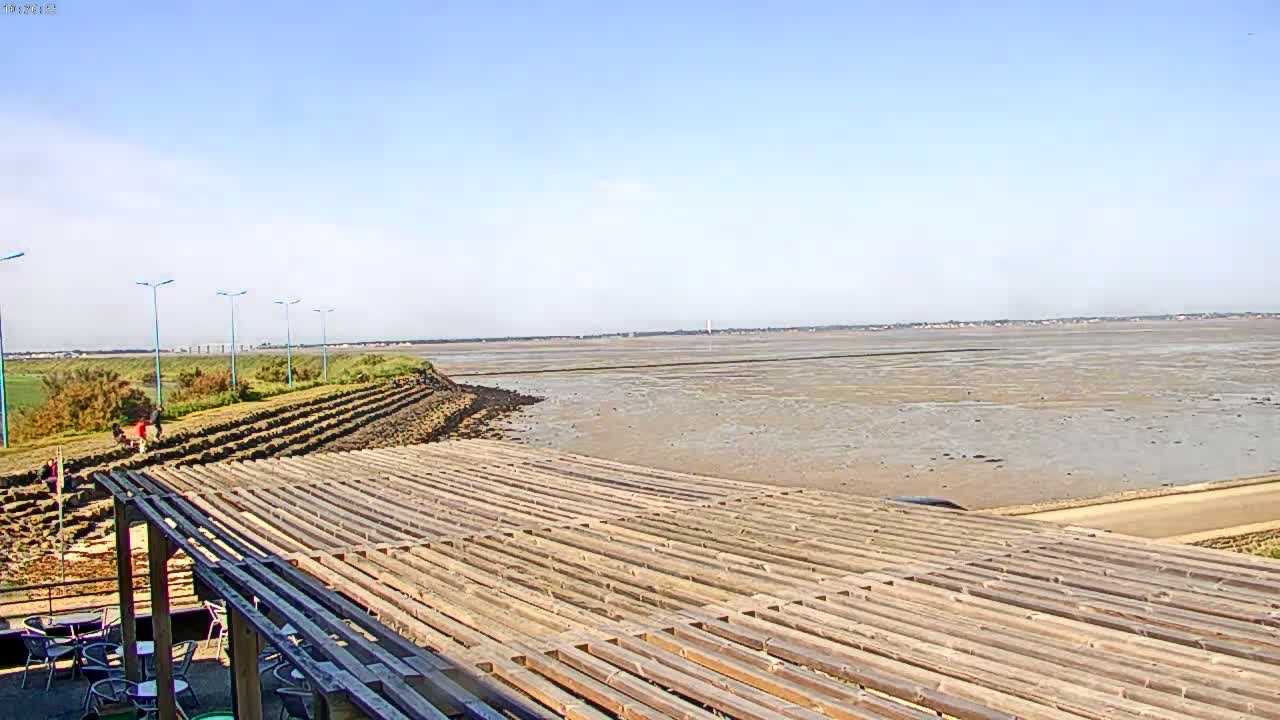 A large expanse of exposed mudflats stretches to the horizon under a clear, sunny sky, overlooked by a wooden pergola and a sparsely populated shoreline.
