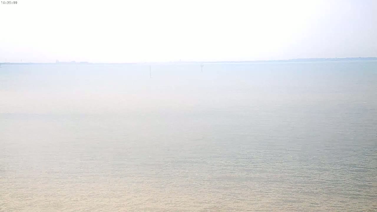A hazy, light-blue expanse of calm water stretches to a distant, indistinct shoreline under a bright, overcast sky.
