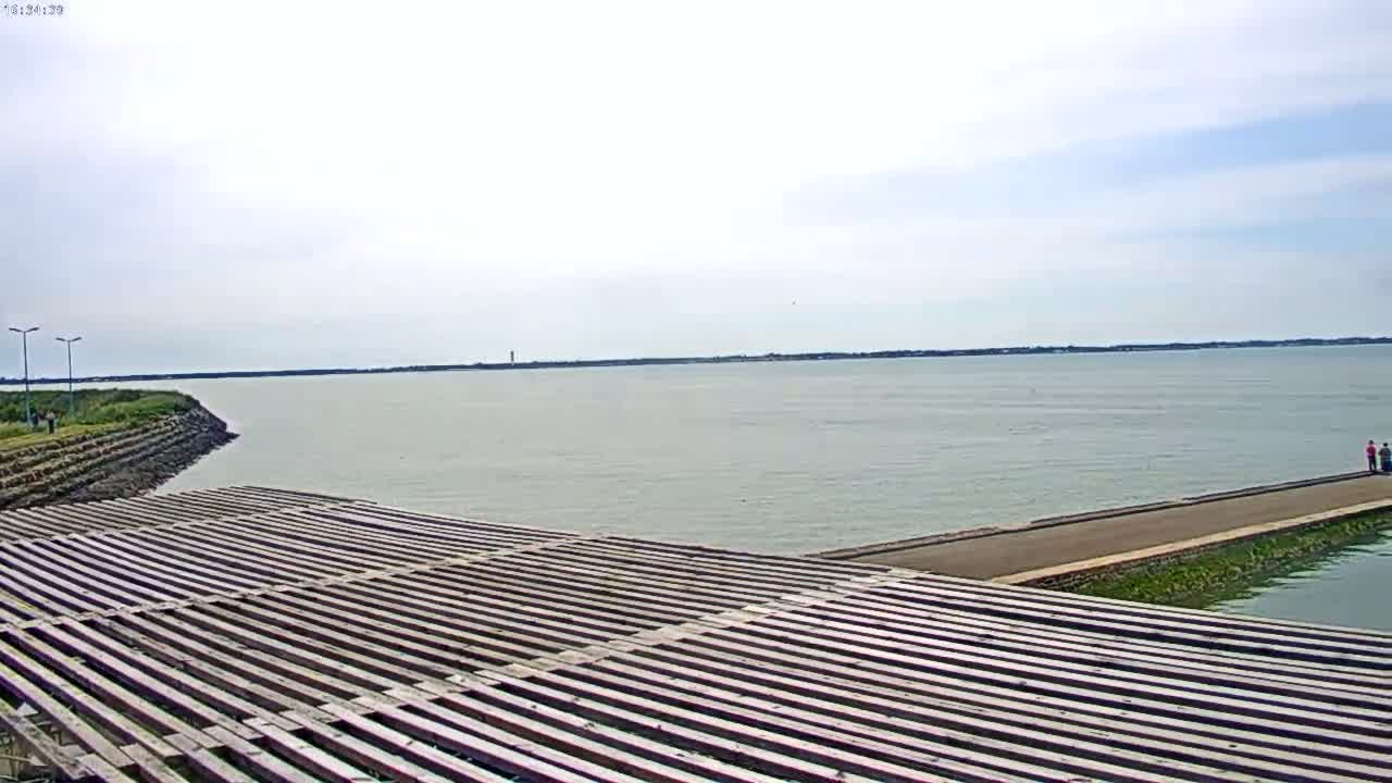 A long wooden structure slopes down to a calm body of water under a mostly cloudy sky.