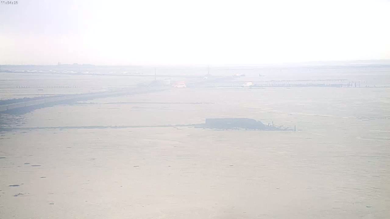 A hazy, flat landscape stretches to the horizon, featuring a long, dark road and a few distant vehicles under a bright, overcast sky.