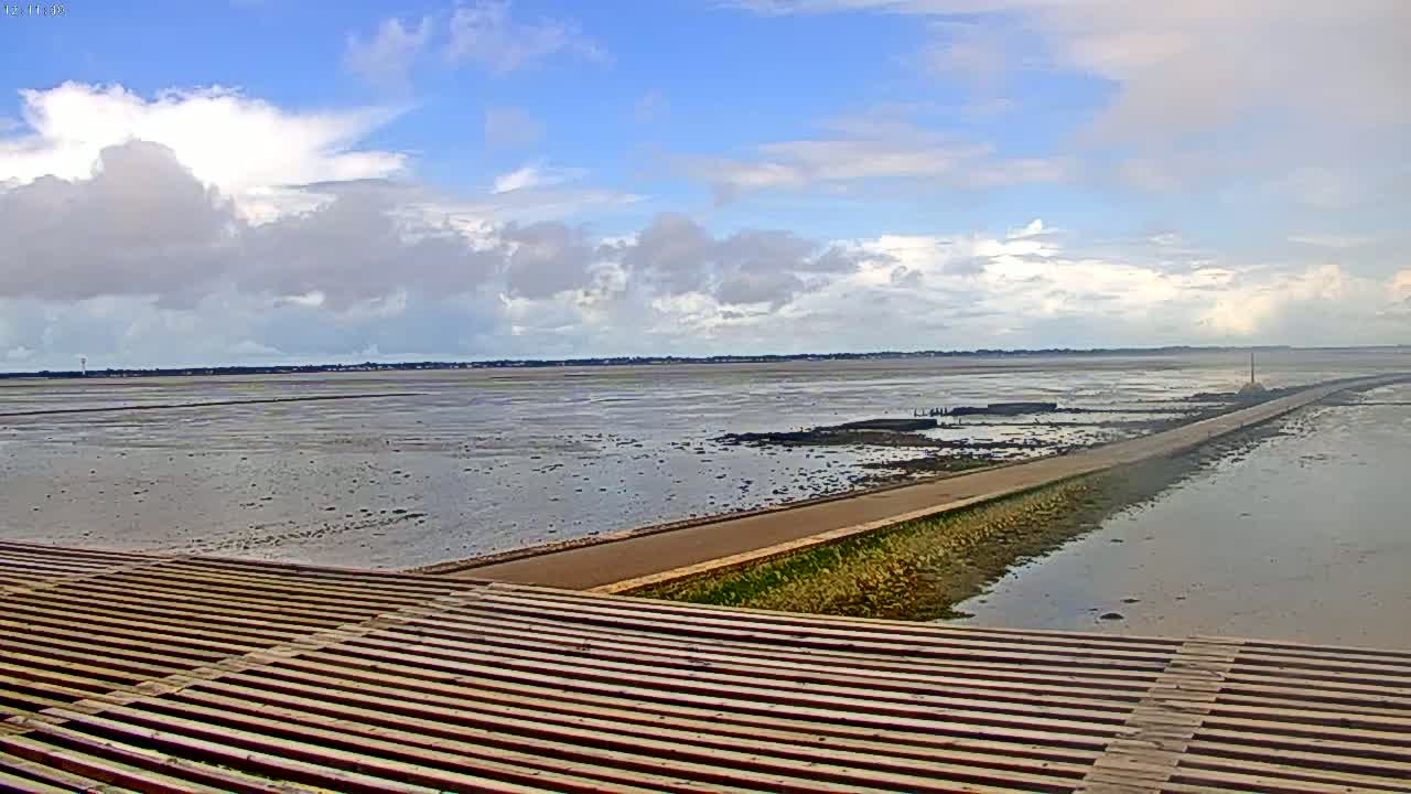 A long, paved causeway extends across a wide expanse of mudflats under a partly cloudy sky.