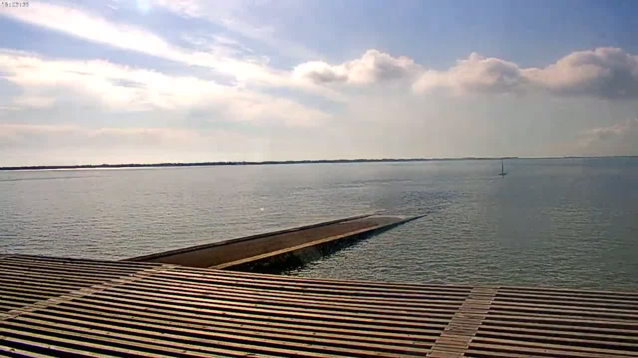 A partly cloudy day reveals a calm body of water with a small sailboat in the distance, viewed from a wooden slatted platform extending over the water.
