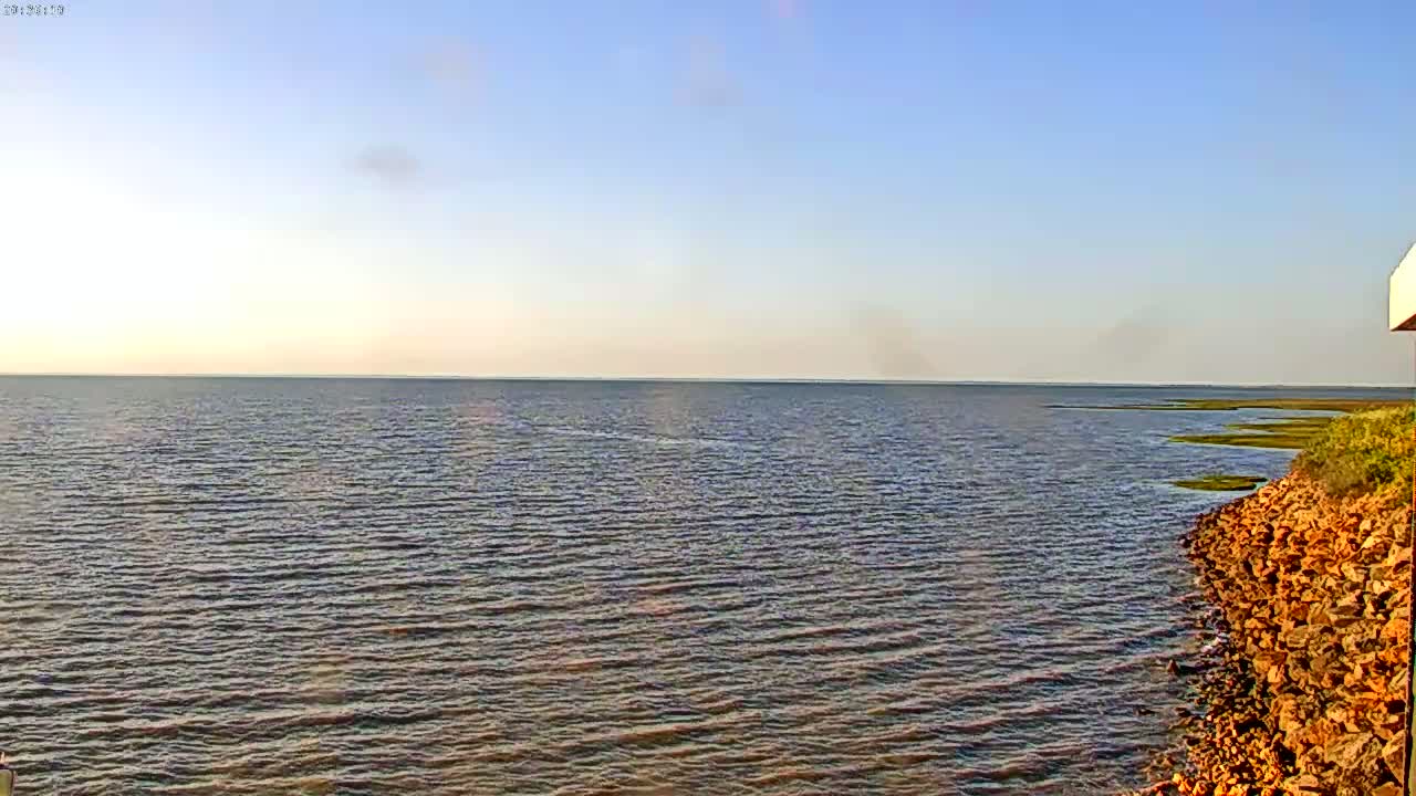 A calm, expansive body of water meets a rocky shoreline under a clear, light blue sky.
