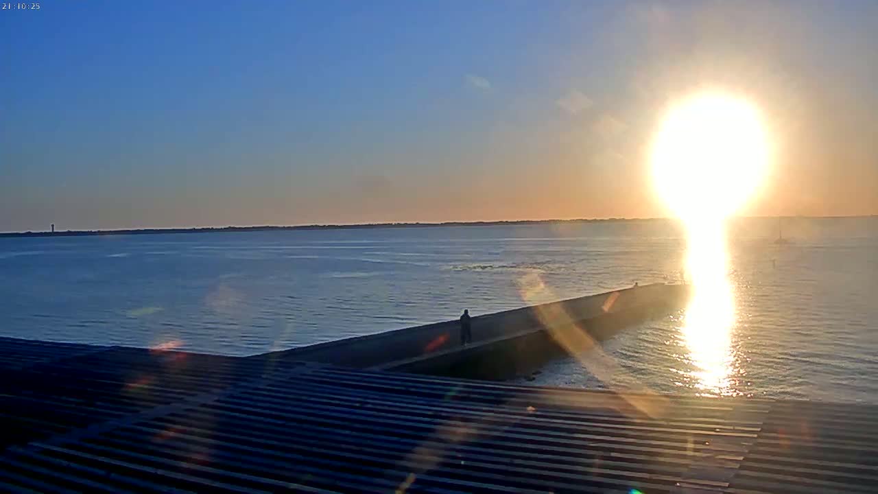 A person stands on a breakwater as the bright sun sets over a calm body of water.