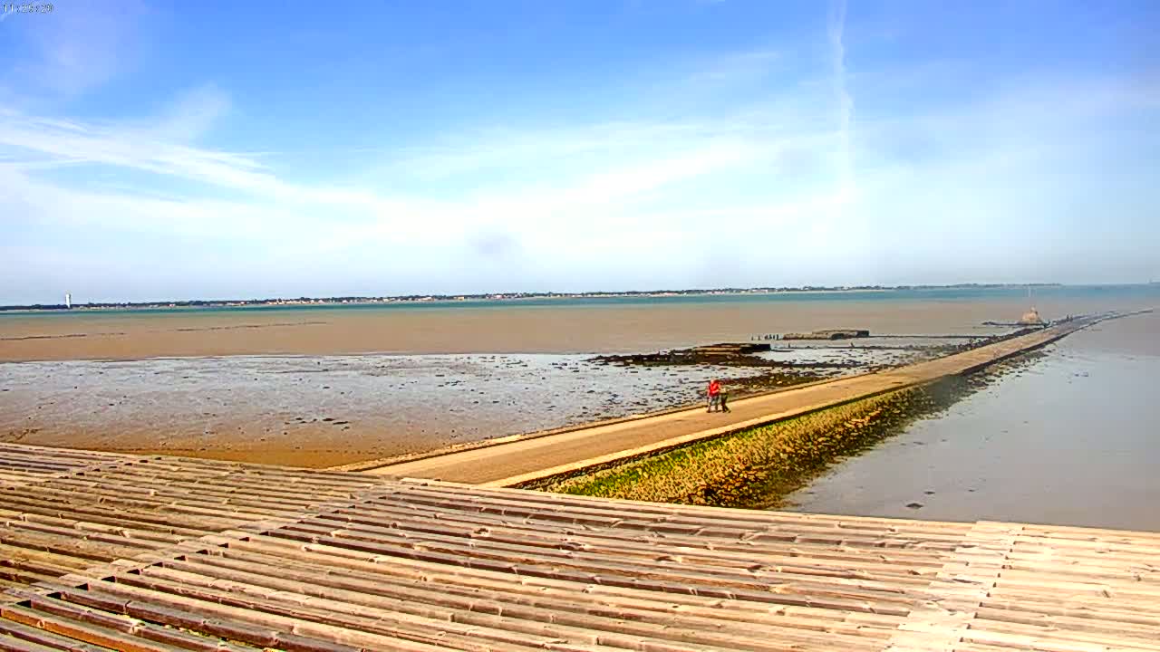 A long, narrow road extends across a wide expanse of shallow, muddy water under a mostly sunny sky.