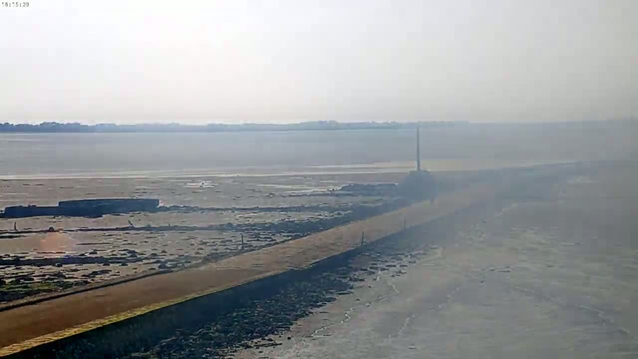 A long, narrow path extends from the foreground across a wide expanse of exposed mudflats and calm, hazy water toward a distant shoreline.