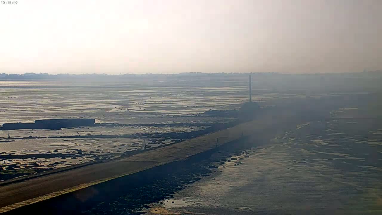 A long, dark road stretches across a hazy, low-tide expanse of water and mudflats toward a distant shoreline under a bright, misty sky.