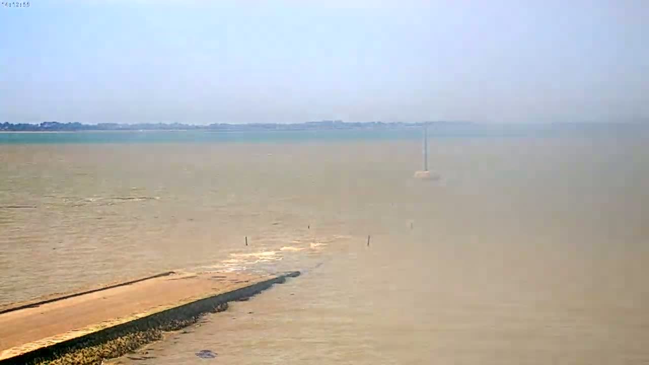 A hazy view shows a long, low concrete structure extending into a calm, light brown body of water, with a distant shoreline visible under a bright, clear sky.