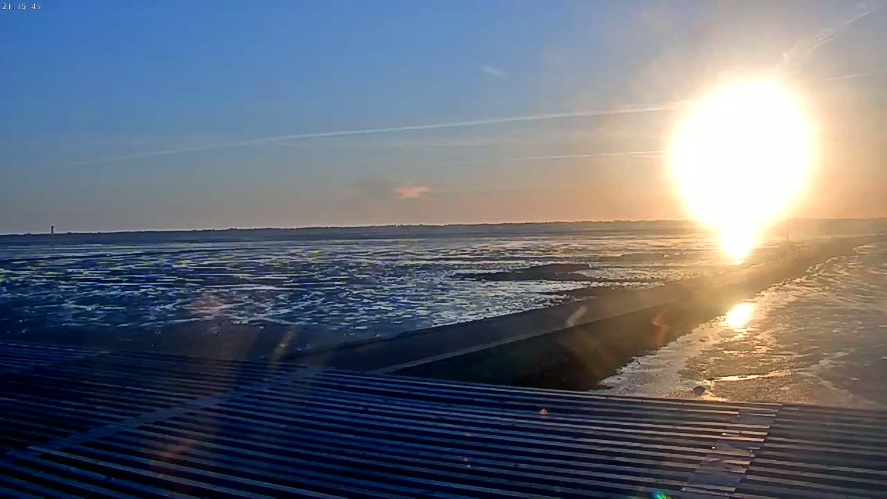 A bright sun sets over a calm, dark-blue body of water, partially reflecting off a paved path, viewed from a ribbed surface.