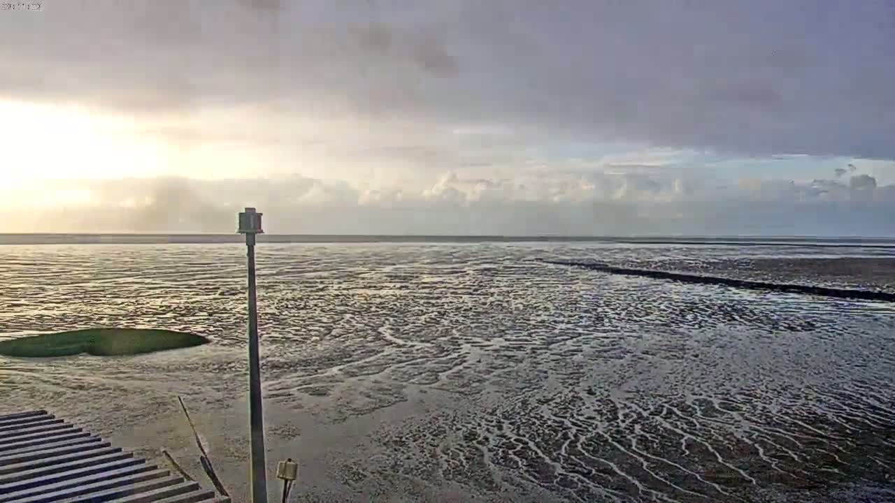 A mostly cloudy sky overlooks a vast expanse of tidal mudflats with shallow water reflecting the light, a single pole stands near a wooden structure.