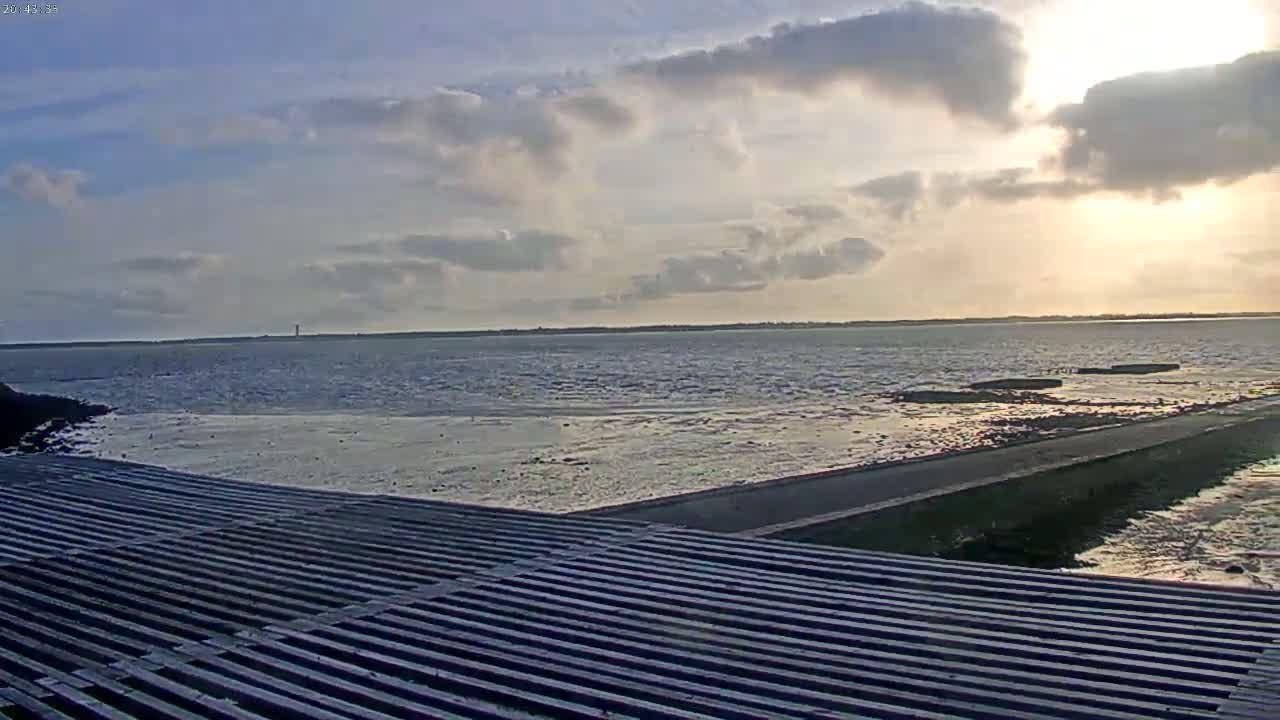 A partly cloudy sunset over a calm body of water, viewed from a slatted wooden surface leading to a paved path.