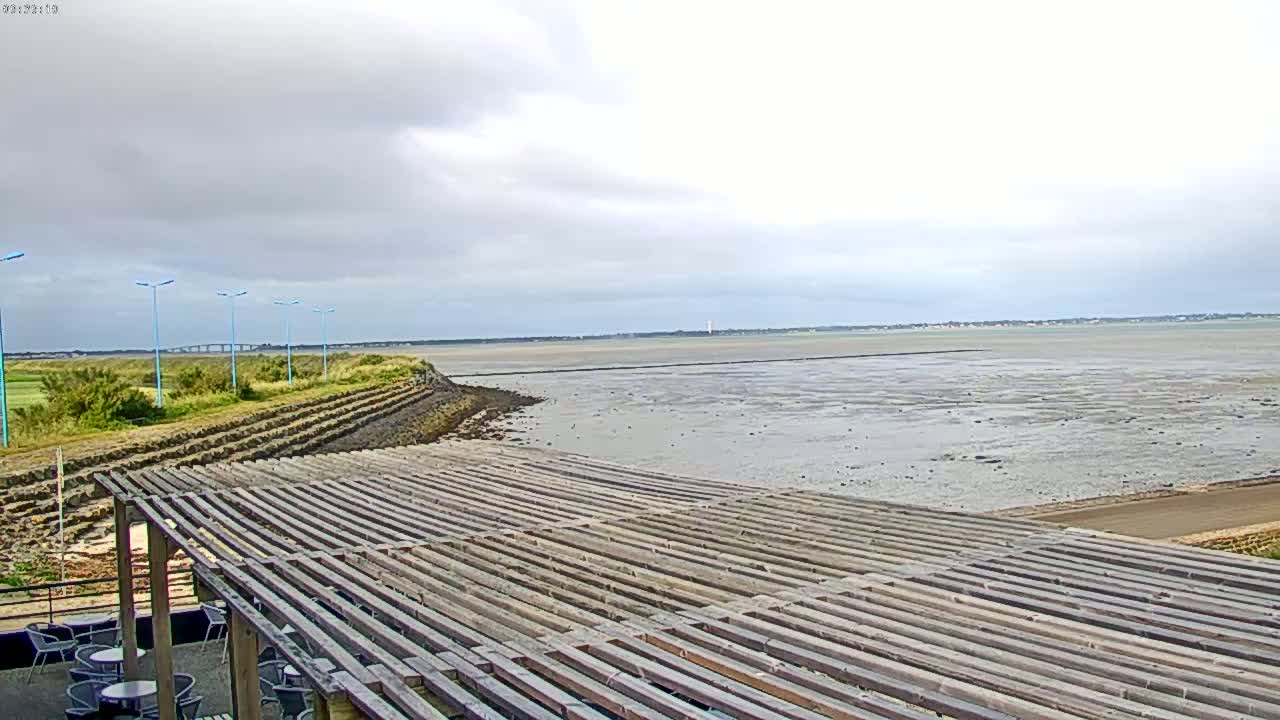 A wooden pergola overlooks a calm, muddy shoreline under a cloudy sky.