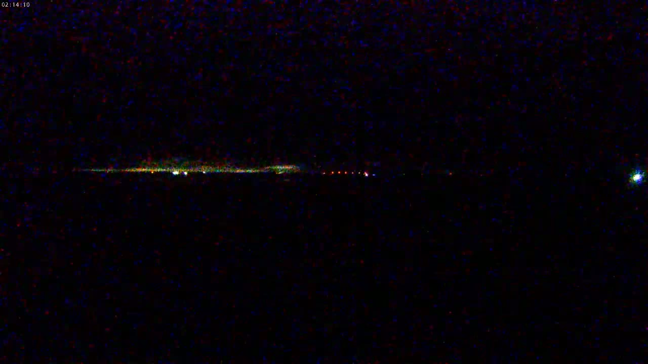 A low-light image shows a long, thin streak of lights in the distance, possibly a distant highway or runway, with a bright light to the upper right, under a dark, nighttime sky.
