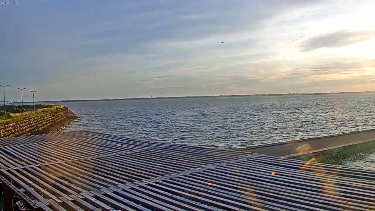 A mostly sunny, slightly breezy day reveals a body of water stretching to the horizon, viewed from a slatted wooden structure near a stone embankment.