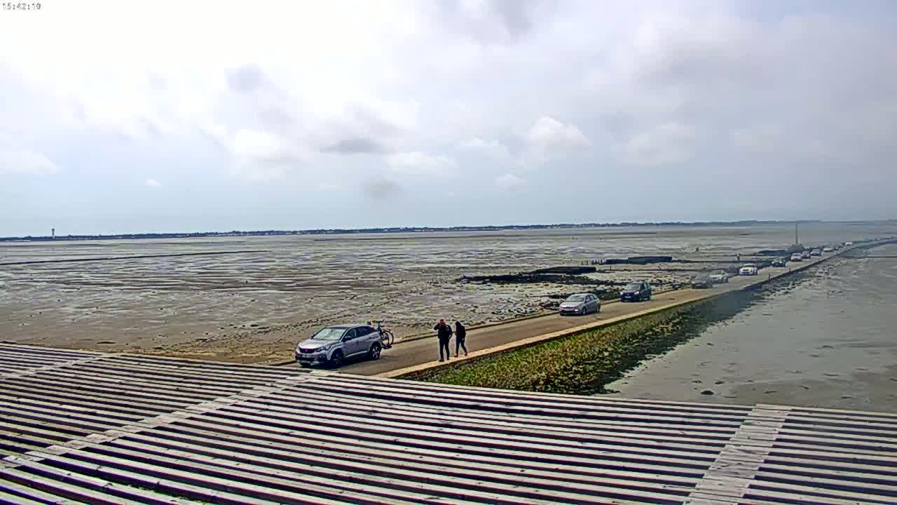 A long road stretches across a wide expanse of mudflats under a partly cloudy sky, with several cars driving along it and people walking nearby.