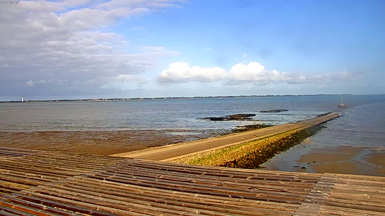 A long, sandy causeway extends into a calm body of water under a partly cloudy sky, with a wooden structure in the foreground.