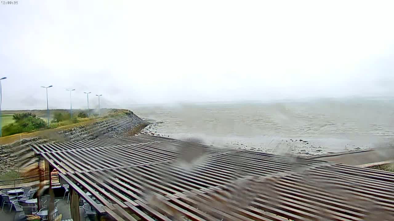 A rain-swept view shows a slatted wooden pergola overlooking a calm, grey sea under an overcast sky.