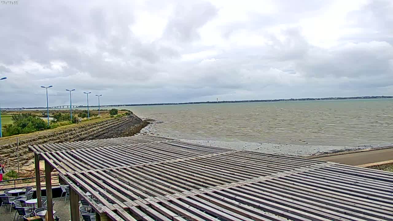 A large wooden pergola sits atop a sloped embankment overlooking a calm, gray body of water under a cloudy sky.