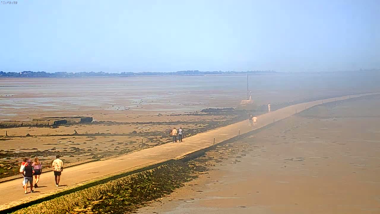 A long, paved path stretches across a hazy, expansive tidal flat toward a distant shoreline under a clear, light-blue sky.