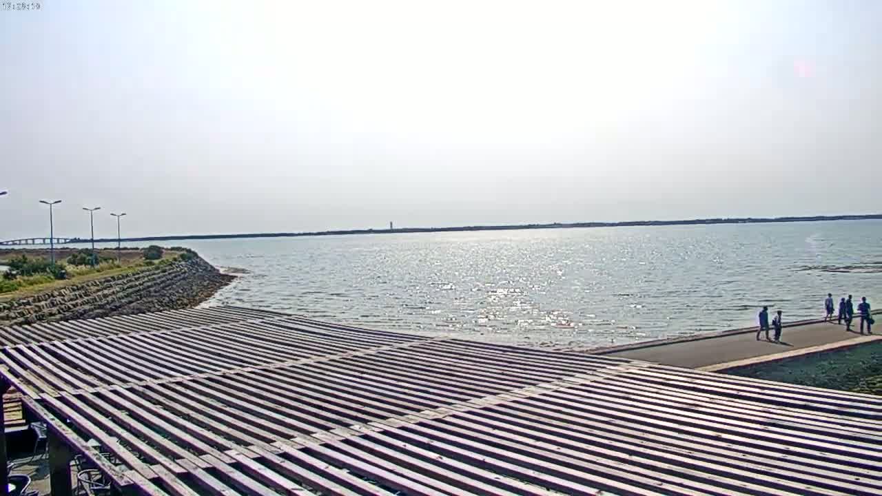 A wooden slatted structure overlooks a calm body of water under a bright, sunny sky, with a few people walking along a paved path in the distance.