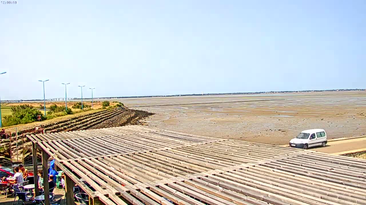 A large, wooden slatted structure sits near a wide expanse of exposed mudflats under a clear, sunny sky, with a single white van visible on a nearby road.