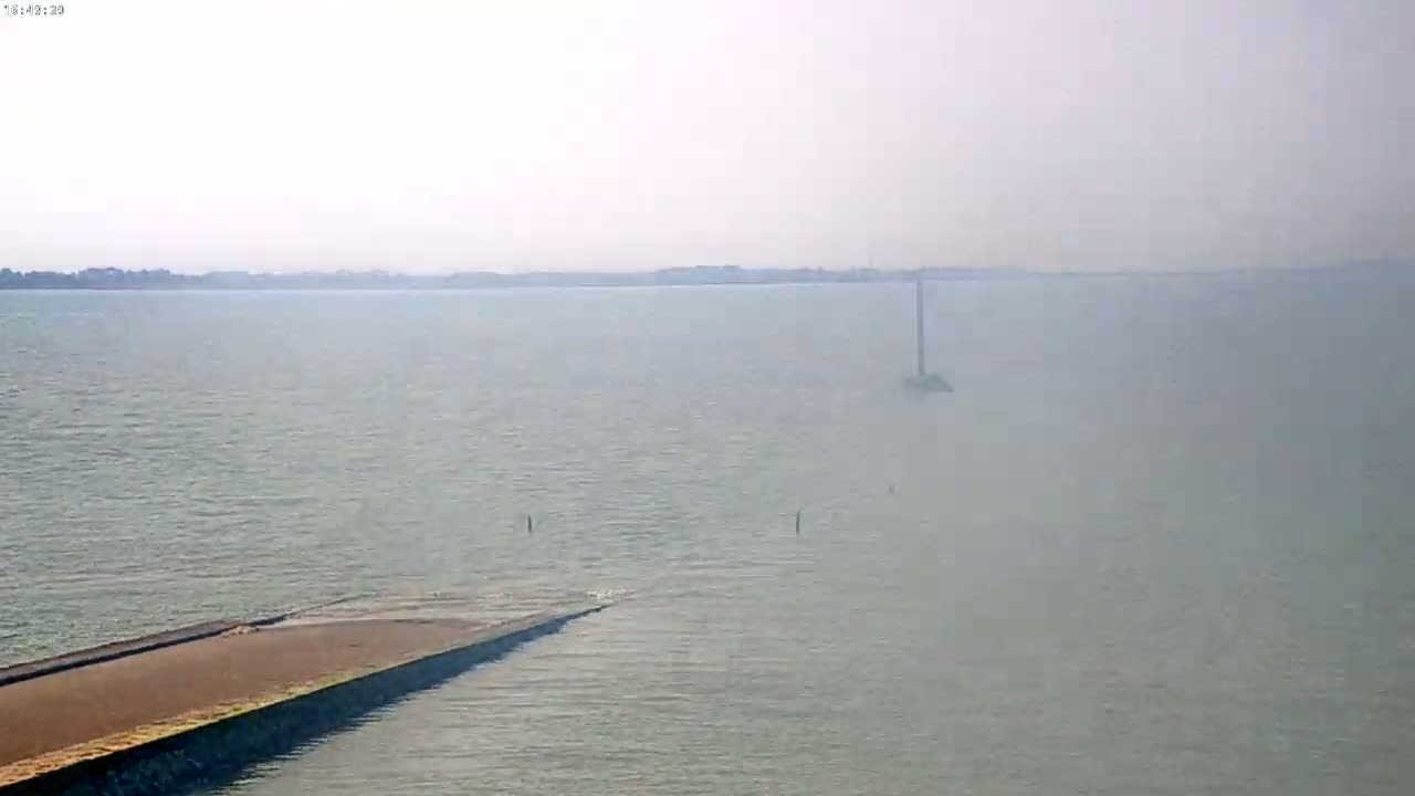 A hazy view shows a calm body of water with a distant shoreline, a tall pole in the water, and a concrete jetty extending from the bottom of the image.