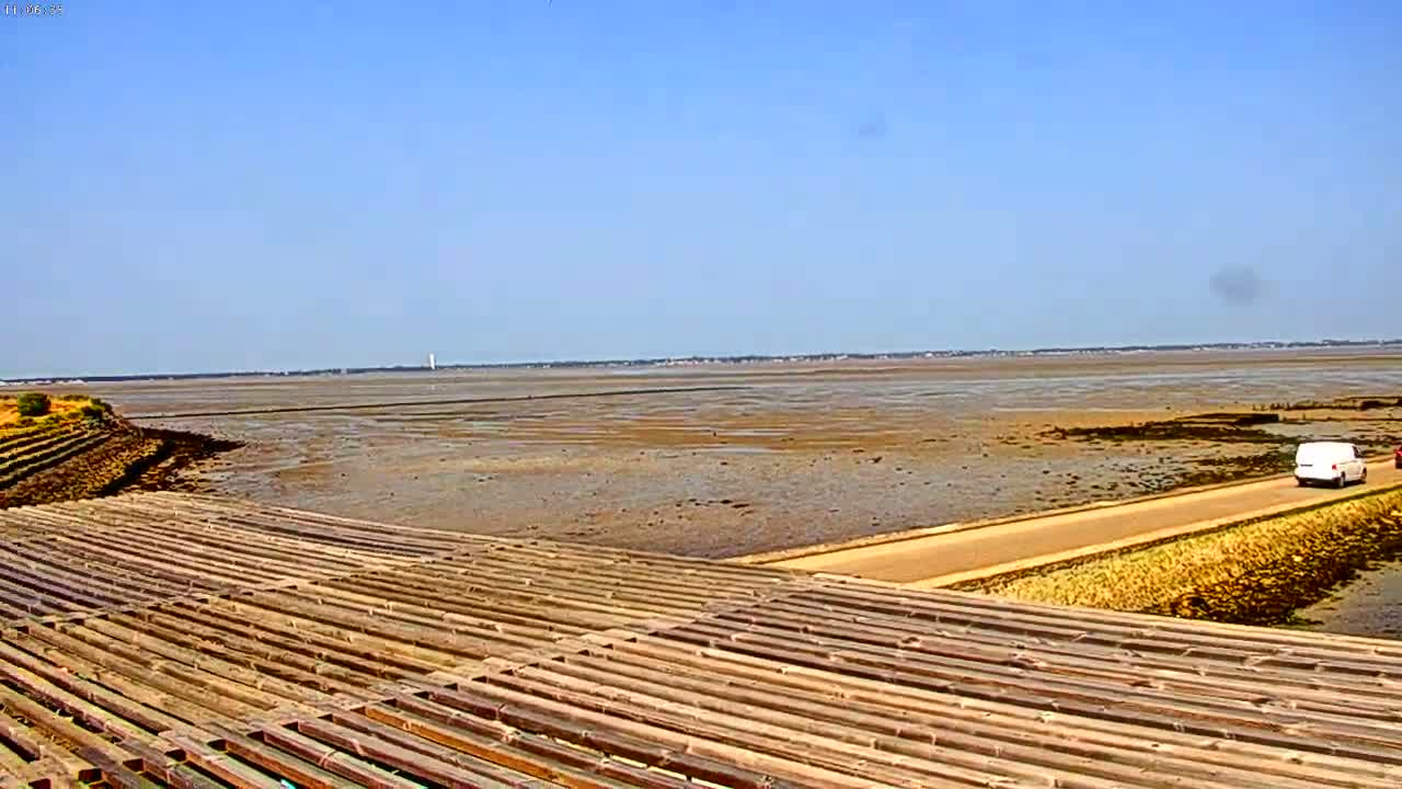 A wide expanse of exposed mudflats stretches to a distant shoreline under a clear blue sky, with a road and a wooden structure in the foreground.
