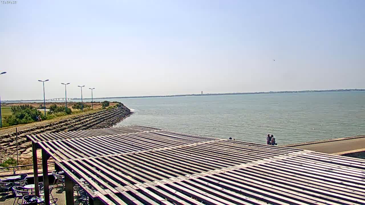 A wooden pergola overlooks a calm body of water under a clear, sunny sky, with a distant bridge visible in the background.