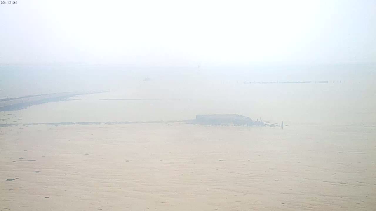 A hazy, foggy view of a flat, pale expanse of water and sand, with a few indistinct structures visible in the middle distance.