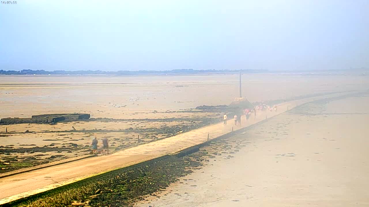 A long, light-colored path stretches across a hazy, expansive mudflat toward a distant shoreline under a mostly clear sky.
