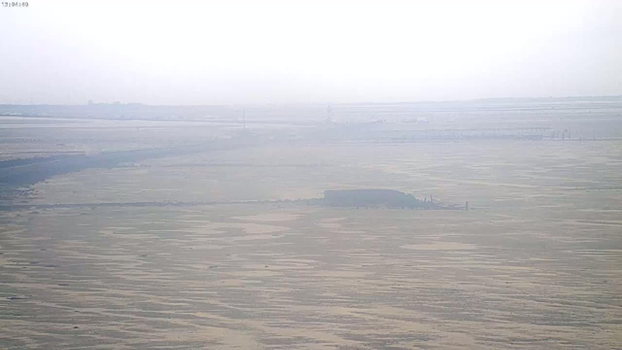 A hazy, expansive flat landscape of sand and water stretches to a distant horizon under a light overcast sky.