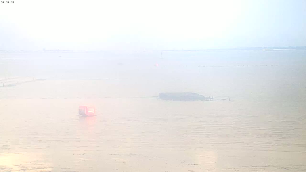 A hazy, light-colored body of water is shown with a small, red object and a larger, dark structure partially submerged in the distance.