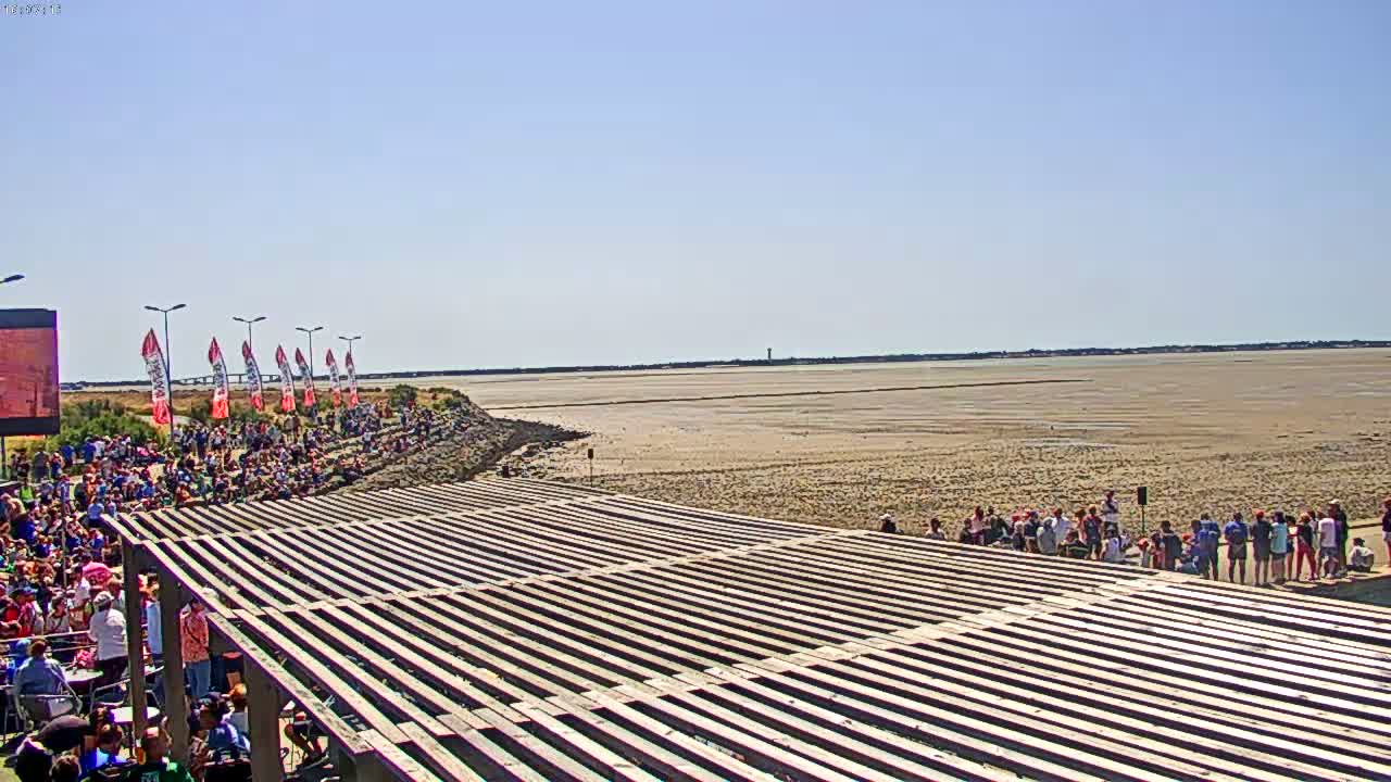 A large crowd of people sits on a sloped viewing area overlooking a wide expanse of exposed mudflats under a clear, sunny sky.