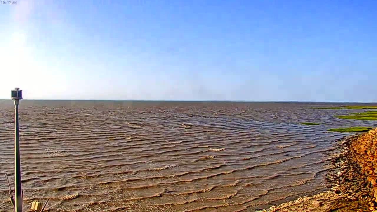 A mostly sunny day shows a body of water with small waves lapping a rocky shoreline under a clear blue sky.